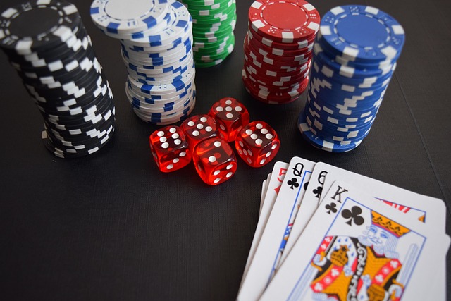 A close-up of poker chips organized in stacks of black, white, blue, green, and red colors, with five red dice showing various numbers. Four playing cards are spread out showing Queen of Clubs, King of Clubs, King of Hearts, and King of Diamonds.