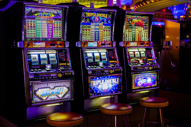 Three colorful slot machines are lit up in a casino. Each slot machine has a unique theme and bright, flashing lights. Three empty stools are positioned in front of the machines, and in the background, more colorful lights and reflections can be seen.