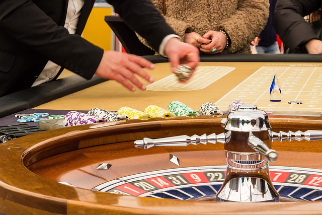 A person is placing chips on a betting table at a casino. The roulette wheel in the foreground shows numbers and pockets. Several other players' hands are visible in the background, and the table is filled with colorful chips.